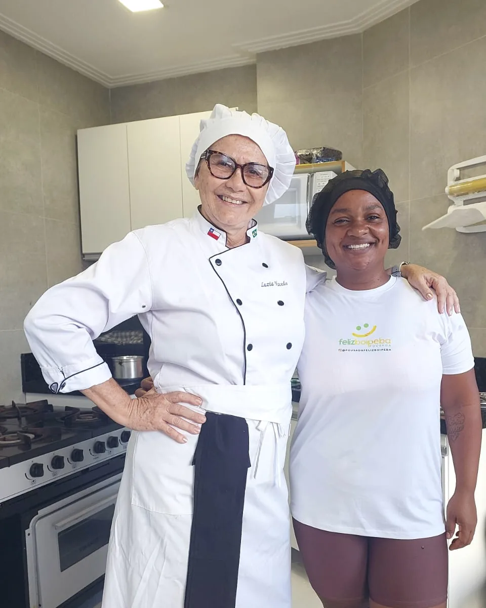 Luzia Macedo, de dólmã branco, ao lado de uma integrante da equipe da cozinha.