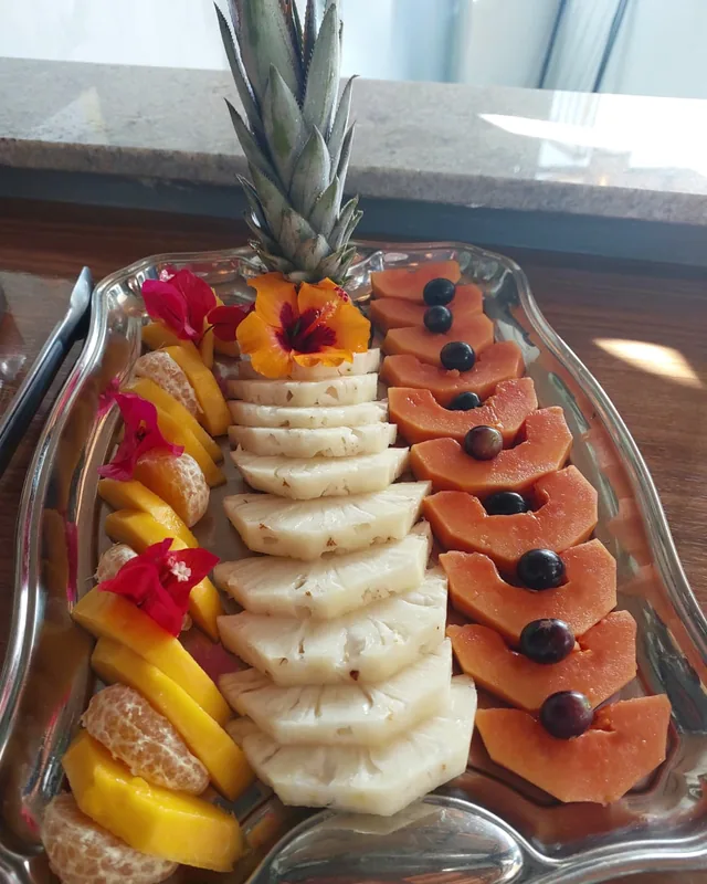 Bandeja de frutas com abacaxi fatiado e sua coroa, mamão com uvas, manga, tangerina e flores de hibisco.