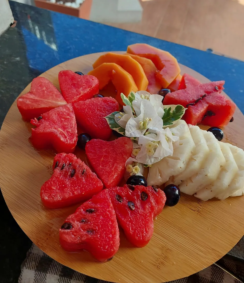 Tábua de frutas com melancia cortada em corações, mamão, abacaxi, uvas e flores brancas, na luz da manhã.