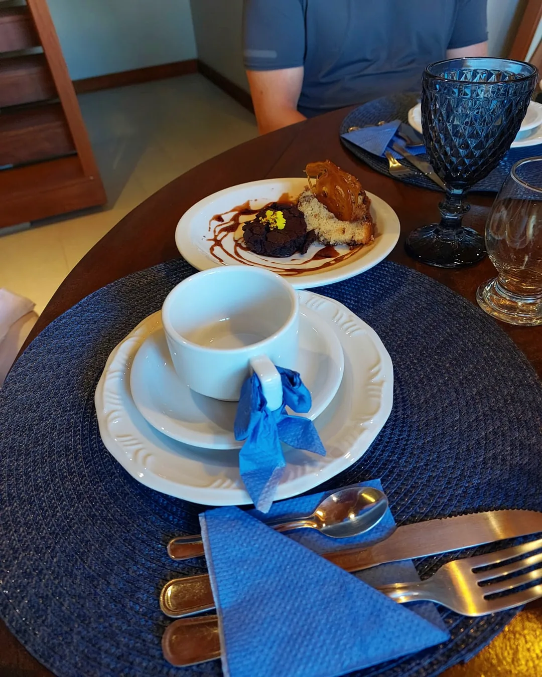 Lugar posto com sousplat azul, xícara com guardanapo amarrado na alça e prato de bolos, com o hóspede à mesa.